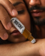 mão de homem aplicando perfume no antebraço de mulher com frasco cilíndrico feito com vidro âmbar e ponta aplicadora com esfera de aço deslizante em primeiro plano e ao fundo rostos juntos do casal